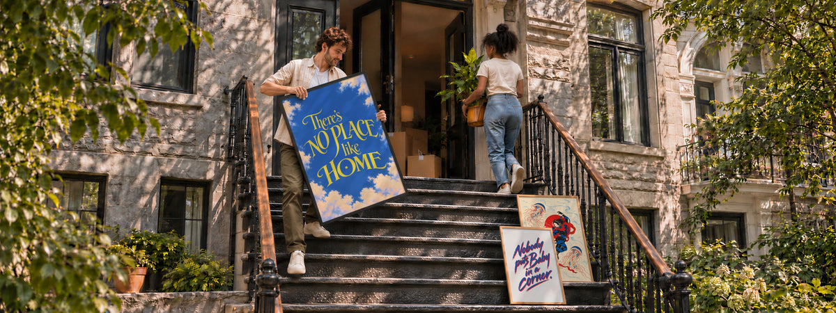A couple moving into a stone townhouse carries a framed print and a plant up the front steps, with two more frames leaning against the railing.
