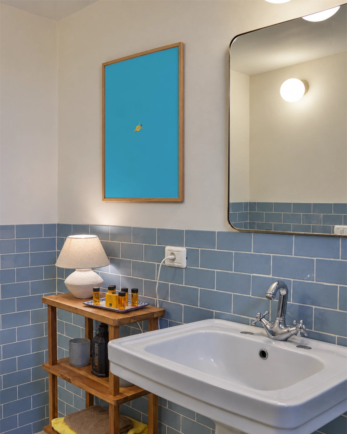 Out of Office art print framed above a wooden shelf in a bathroom with pale blue tiles, featuring a tiny swimmer on a yellow ring.