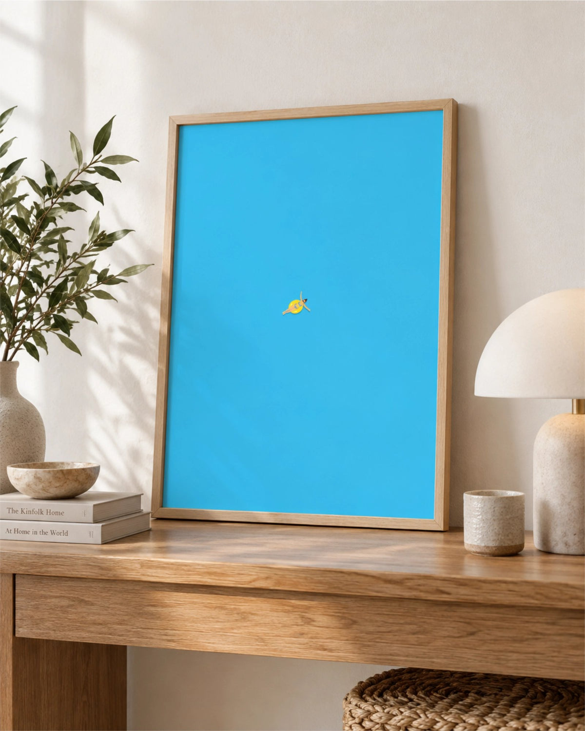 Out of Office art print in a natural wood frame leaning on a wooden console table, showing a tiny swimmer floating in bright blue water.