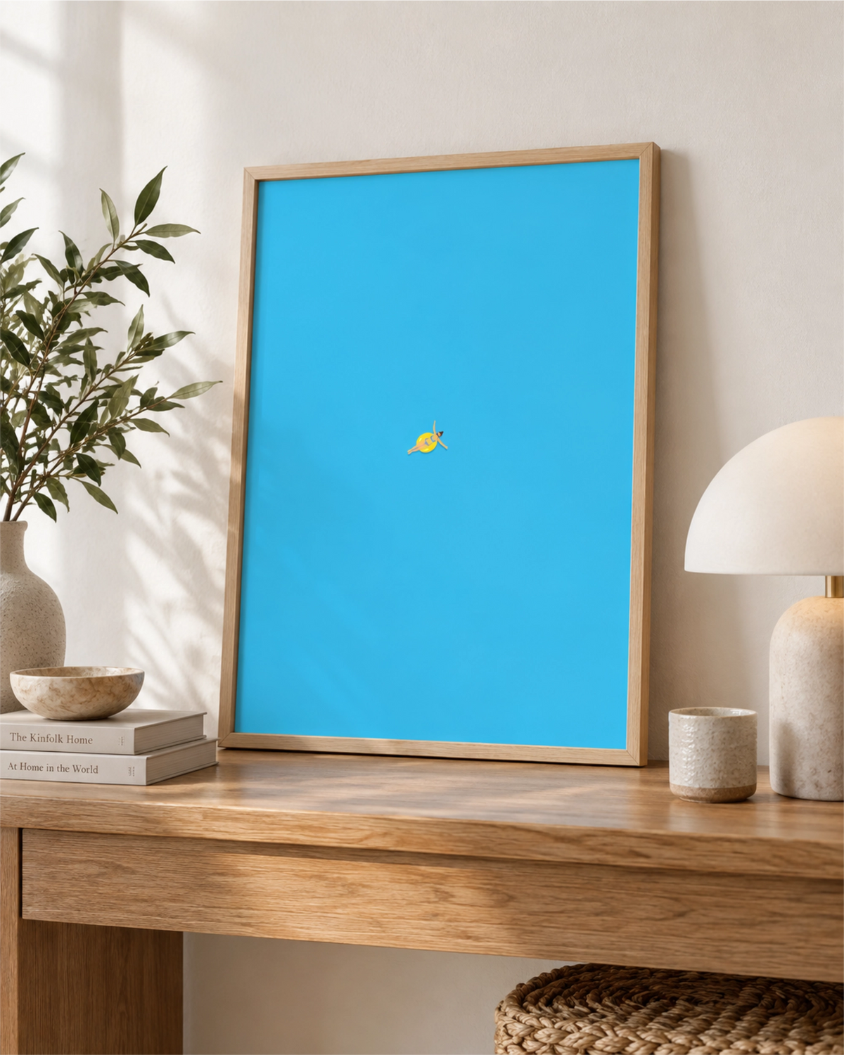 Out of Office art print in a natural wood frame leaning on a wooden console table, showing a tiny swimmer floating in bright blue water.