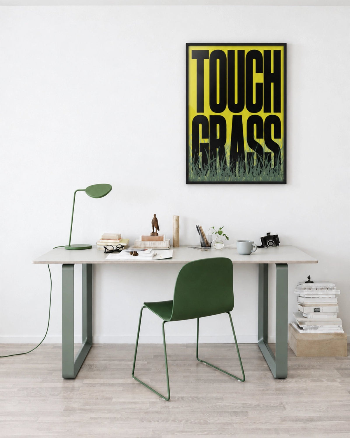 Affiche Touch Grass en cadre noir au-dessus d’un bureau blanc dans un bureau à domicile épuré avec chaise verte et lampe assortie