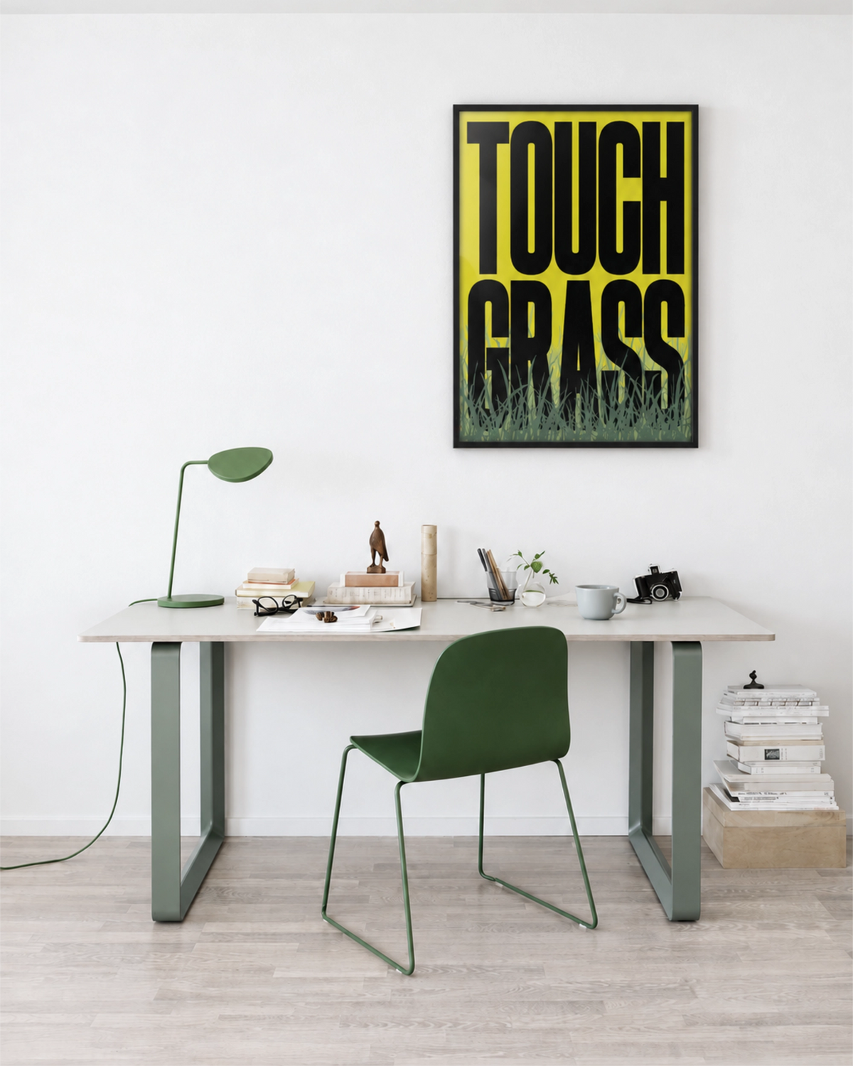 Touch Grass art print in a black frame above a white desk in a minimalist home office with a green chair and matching desk lamp.