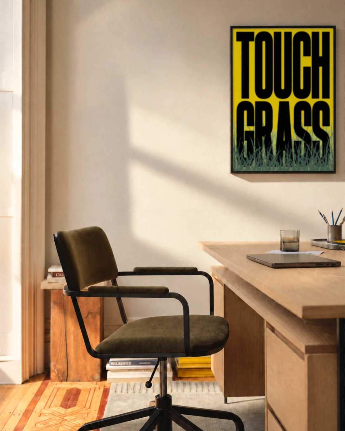 Affiche Touch Grass encadrée au-dessus d’un bureau en bois dans un espace de travail ensoleillé, typographie noire sur jaune et herbe illustrée
