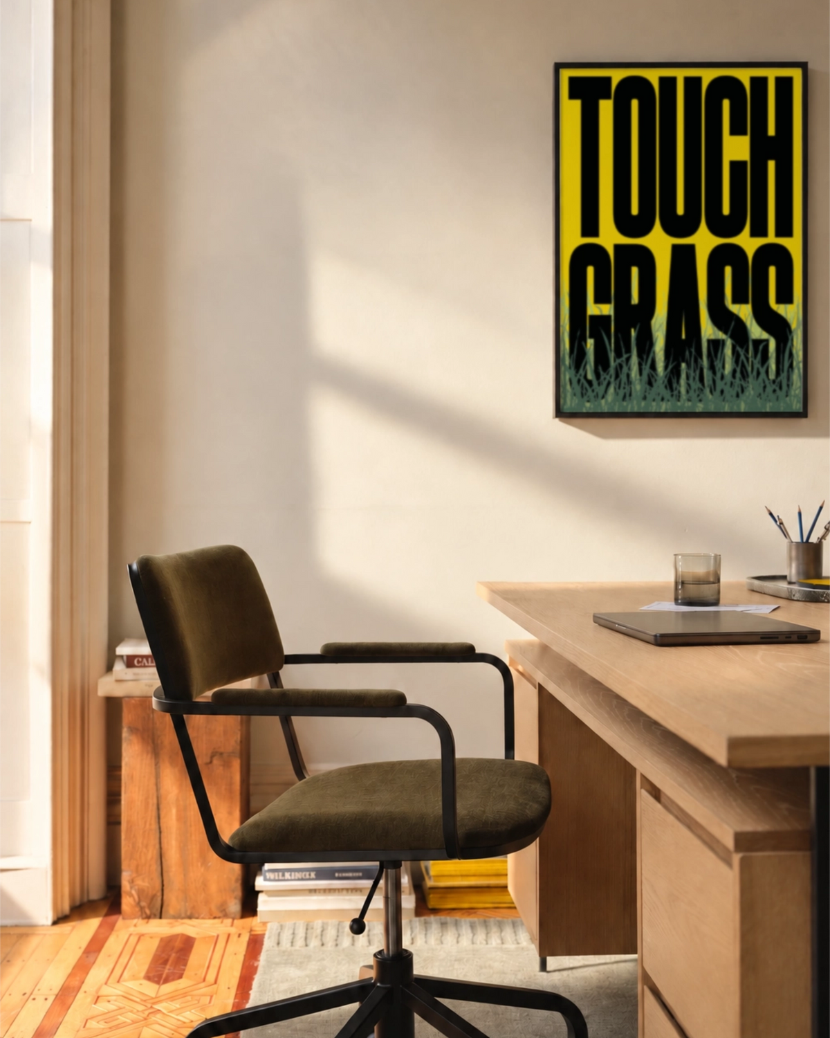 Touch Grass art print framed above a wooden desk in a sunlit workspace, featuring bold black typography on yellow with illustrated grass.