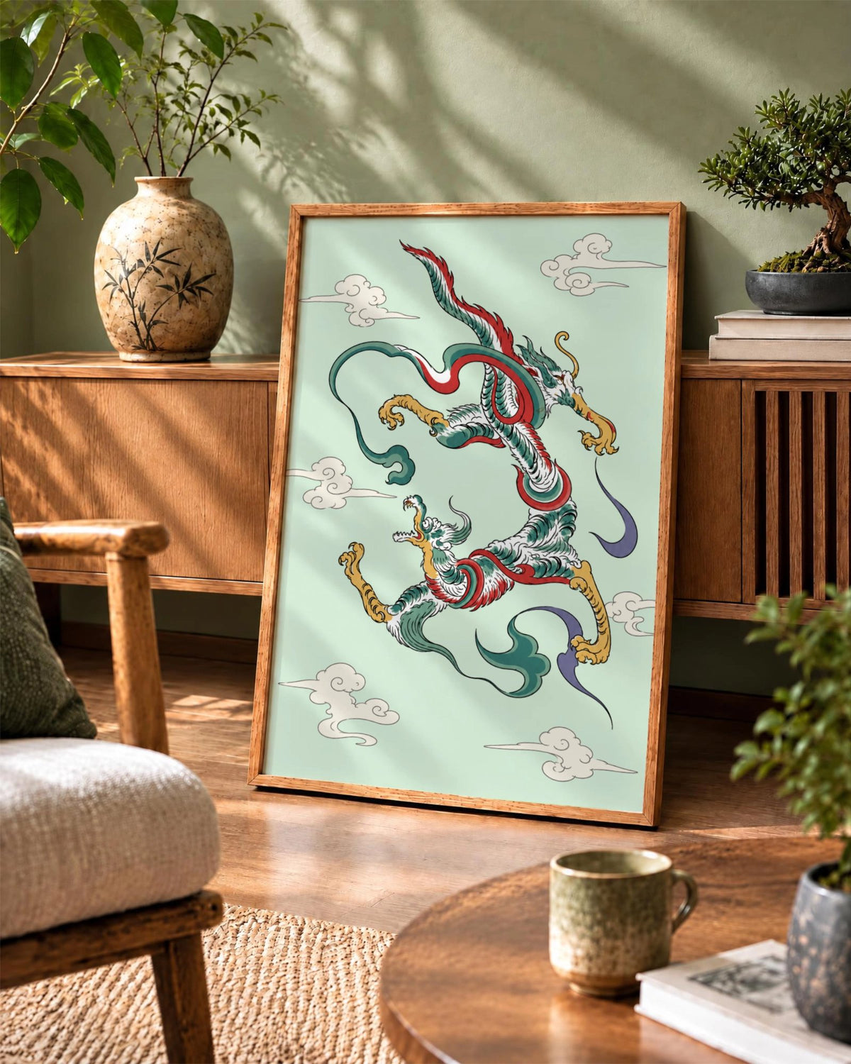 Dragon Dance art print in a natural wood frame leaning against a green wall beside a ceramic vase, bonsai tree and wooden cabinet.