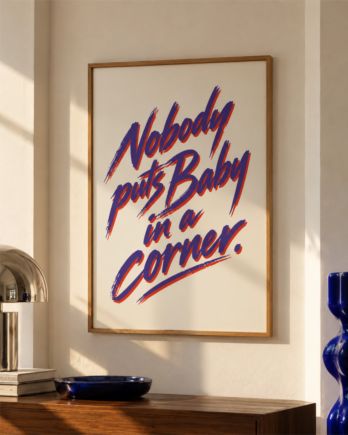 Nobody Puts Baby in a Corner typography art print in a natural wood frame above a dark walnut console, styled with blue ceramics in warm sunlight.