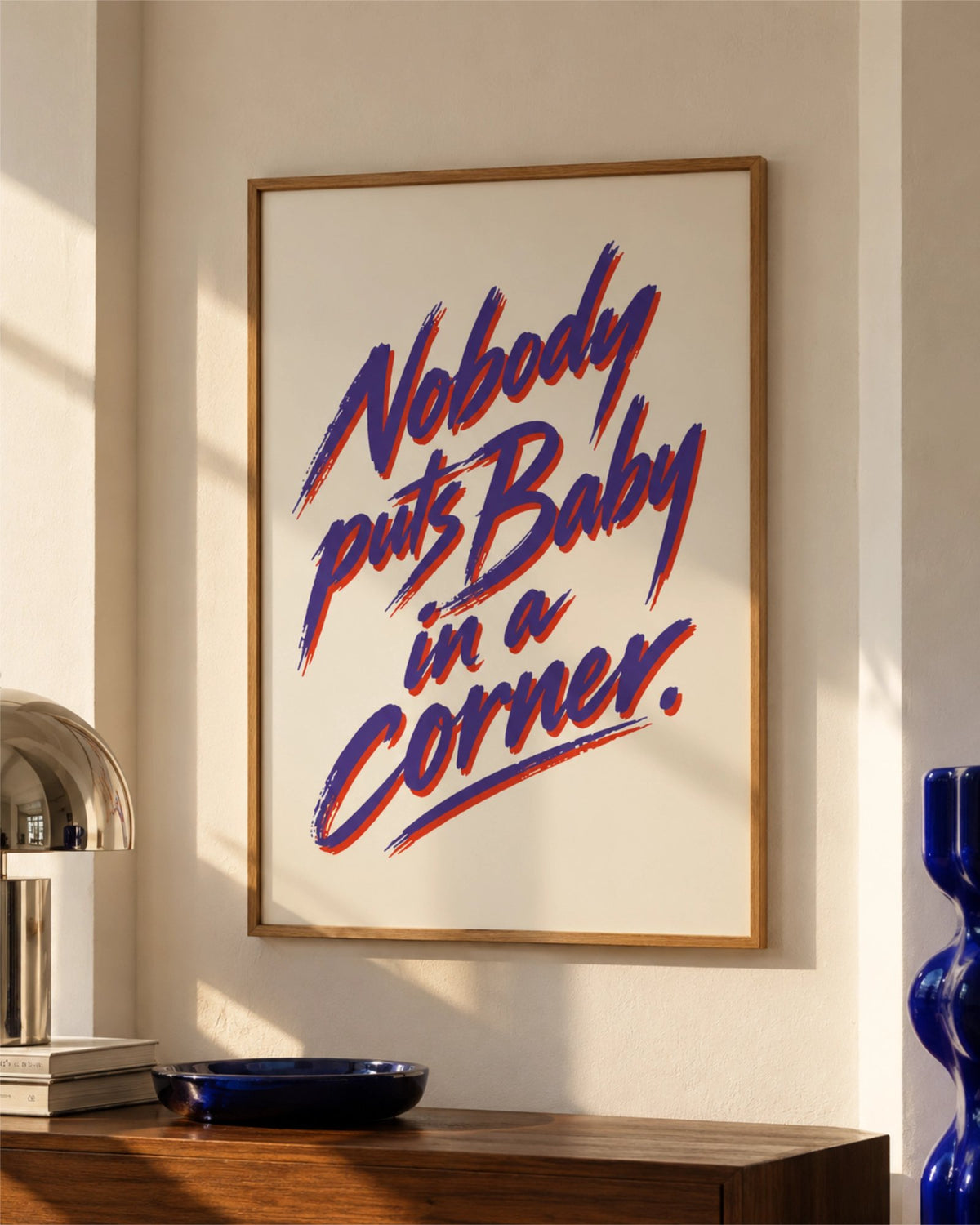 Nobody Puts Baby in a Corner typography art print in a natural wood frame above a dark walnut console, styled with blue ceramics in warm sunlight.