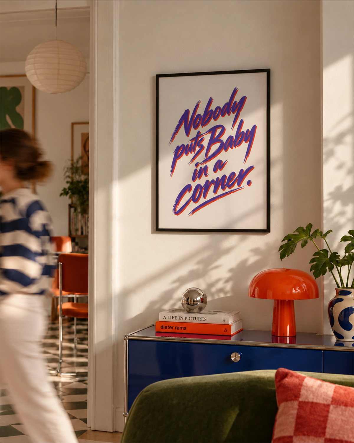 Poster Nobody Puts Baby in a Corner in een zwarte lijst boven een blauwe kast met een oranje paddenstoellamp en een kamerplant, in een kleurrijke, moderne woonkamer.
