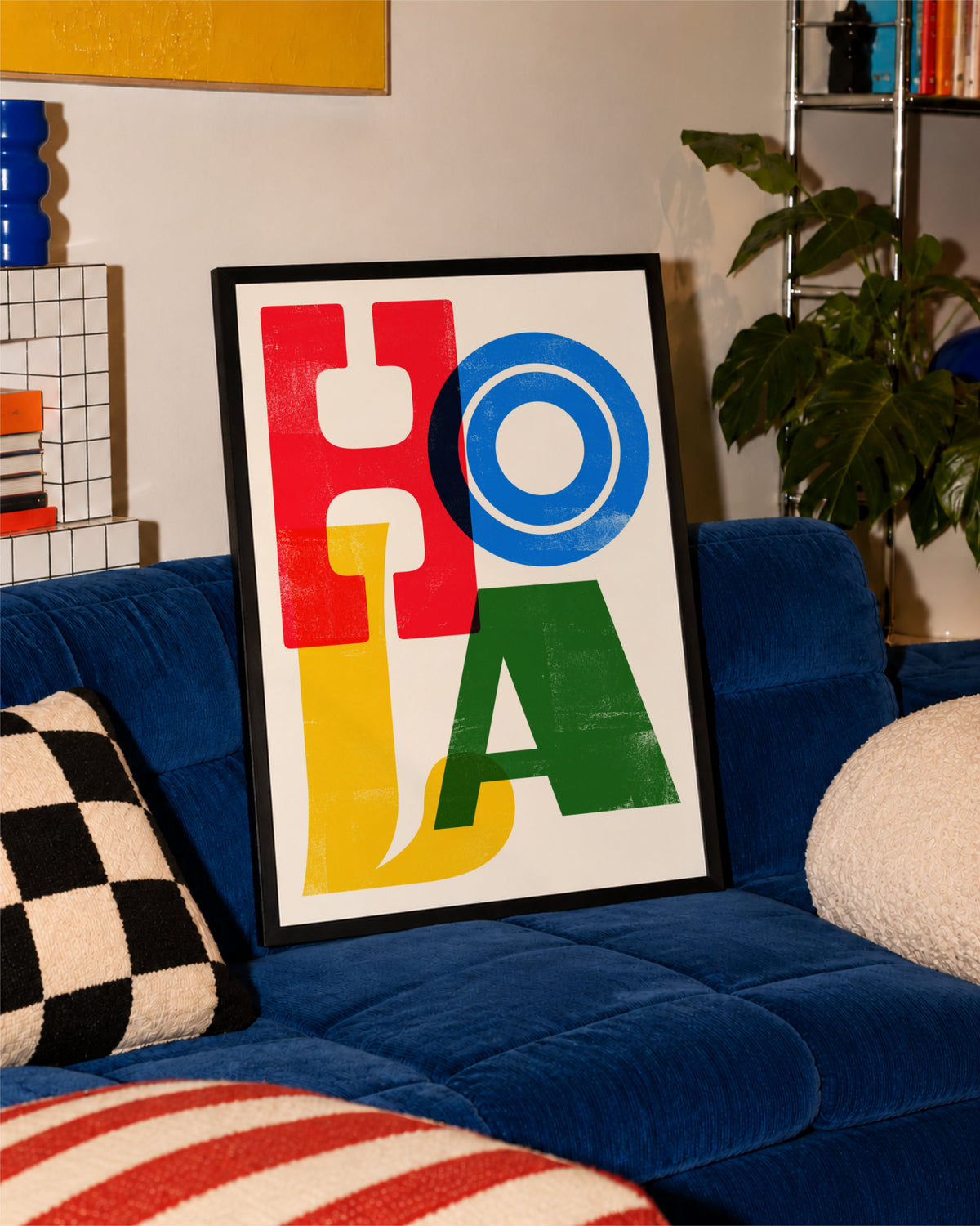 Black-framed Hola poster leaning against a vivid blue sofa with patterned cushions