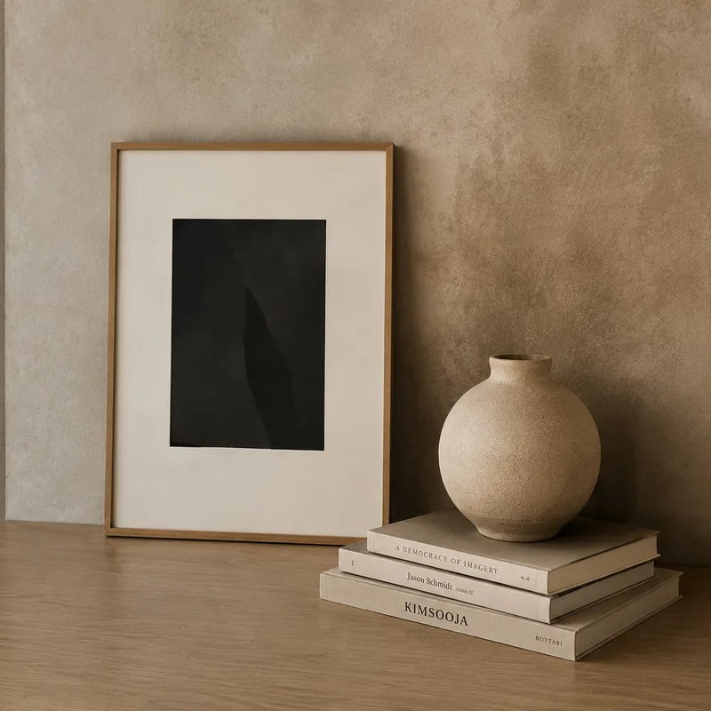 Framed black abstract print leaning on a wooden shelf beside a round ceramic vase and art books